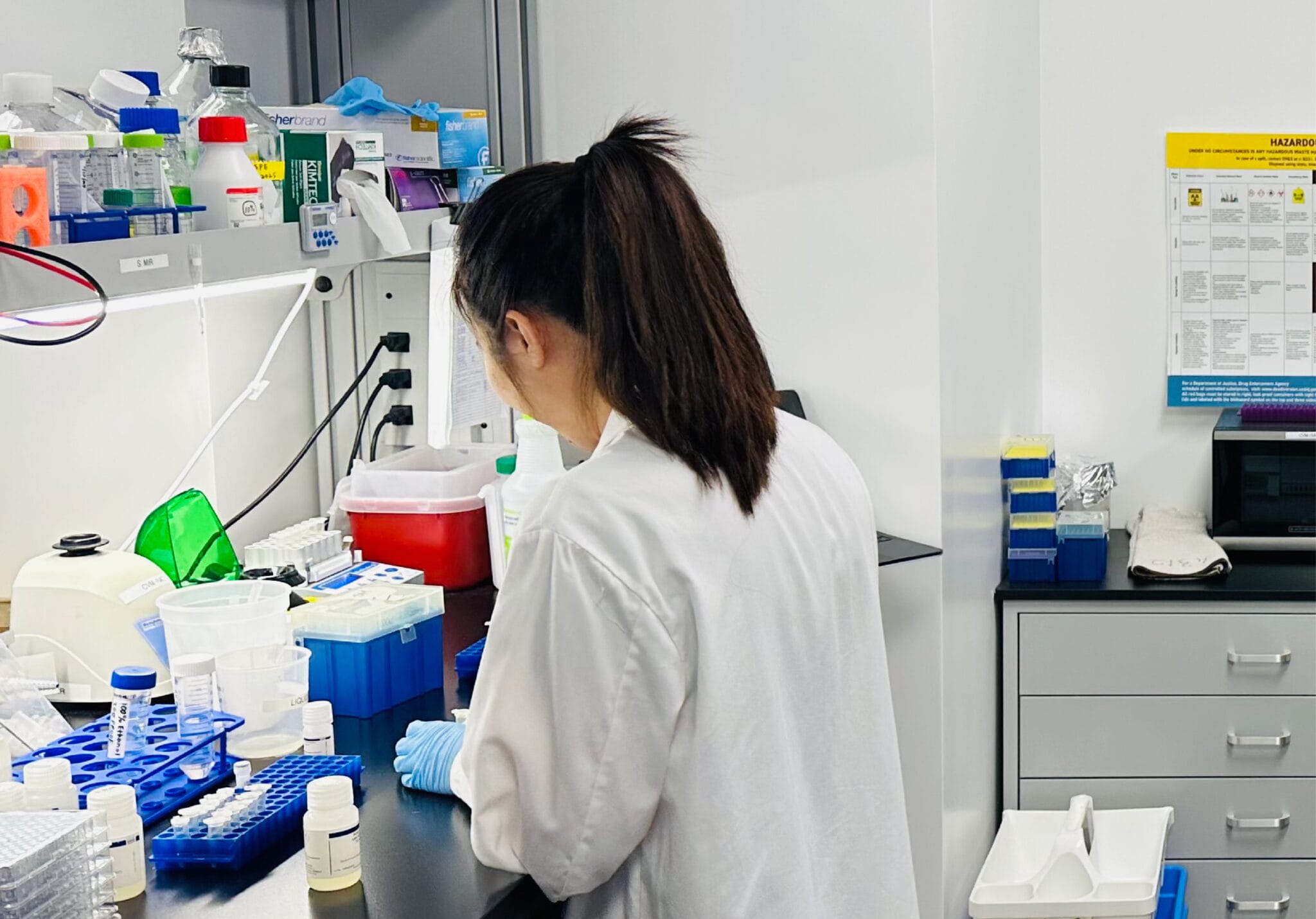 A person wearing a white lab coat and blue gloves works at a laboratory bench with various scientific equipment and supplies.